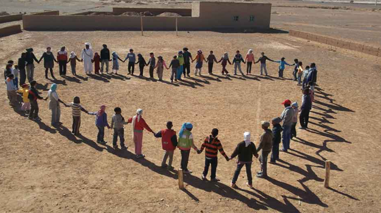 People form heart shape on desert sand with travel-themed text overlays by moroccotripway together creating memorable group moment in Sahara.