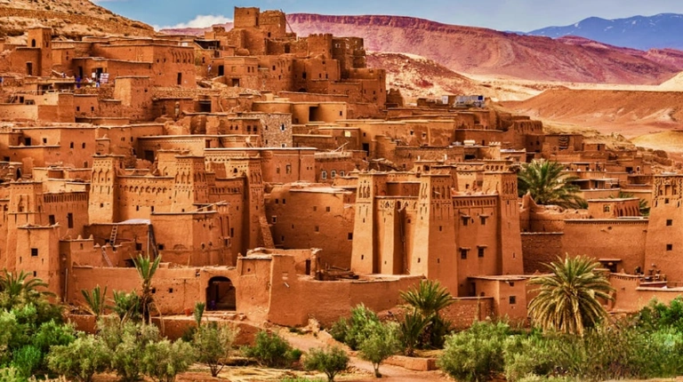 Ancient fortified village rises from desert landscape, featuring earthen towers, narrow passages, and terraced structures beneath a clear sky.