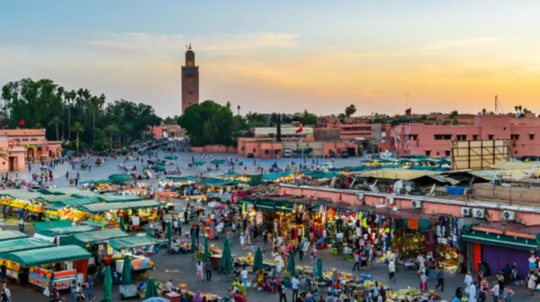 Crowded square glows at sunset with performers, food stalls, and gathering crowds as evening lights begin illuminating the scene.