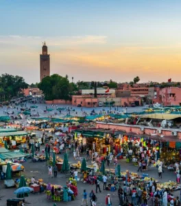 Crowded square glows at sunset with performers, food stalls, and gathering crowds as evening lights begin illuminating the scene.