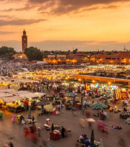 Vibrant city square fills with crowds, market stalls, and performers at sunset, glowing lights emerging as evening activity builds.