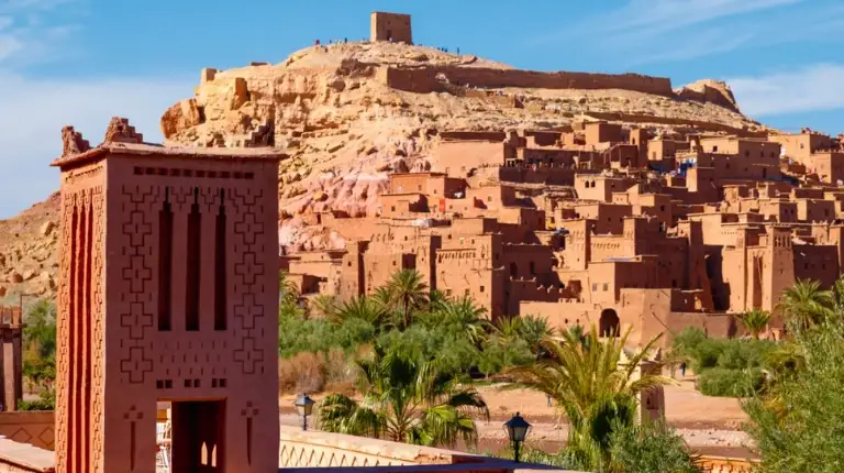 Historic earthen fortified village with towered walls rising along a hillside, showcasing traditional southern Moroccan architecture in sunlit desert landscape.
